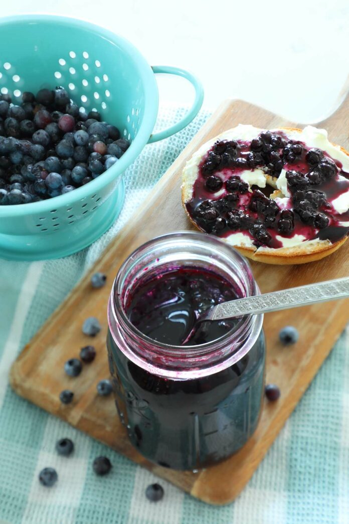 A jar of blueberry jam on a cutting board with fresh blueberries and a bagel.