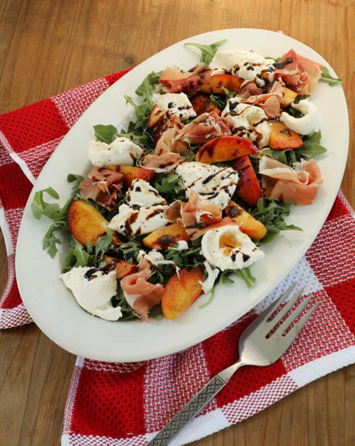 Peach and burrata salad on a white platter sitting with a red kitchen towel and silver fork.