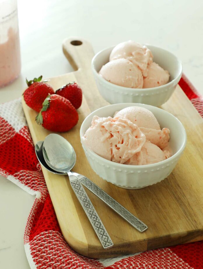 Two bowls of strawberry ice cream on a cutting board with fresh strawberries.