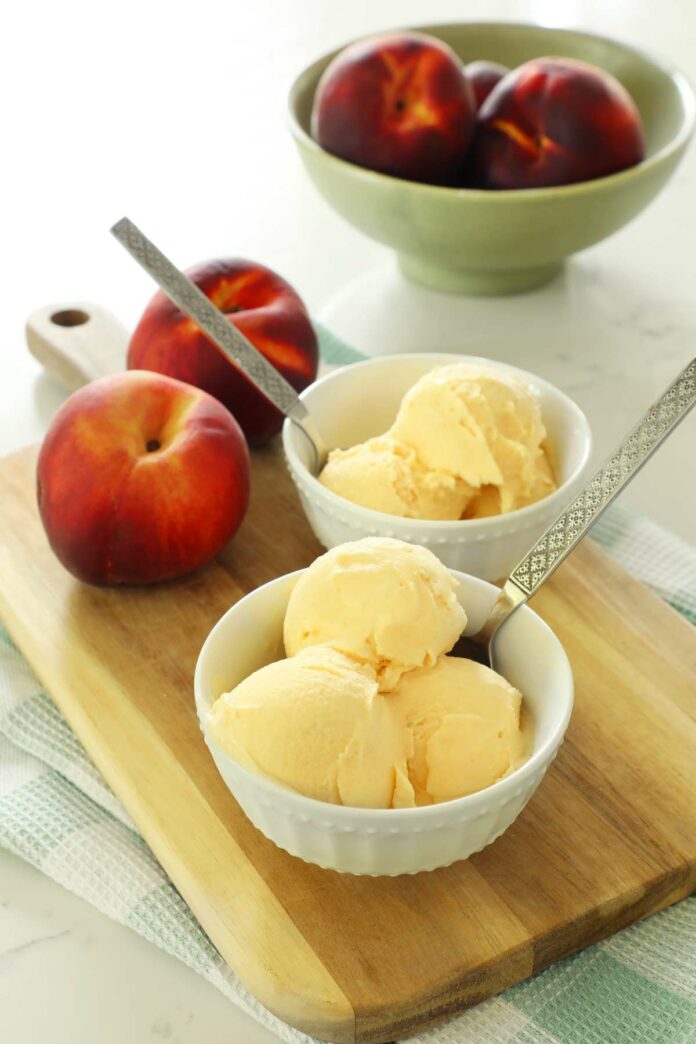 Peach ice cream in two white bowls on a wooden cutting board with fresh peaches.