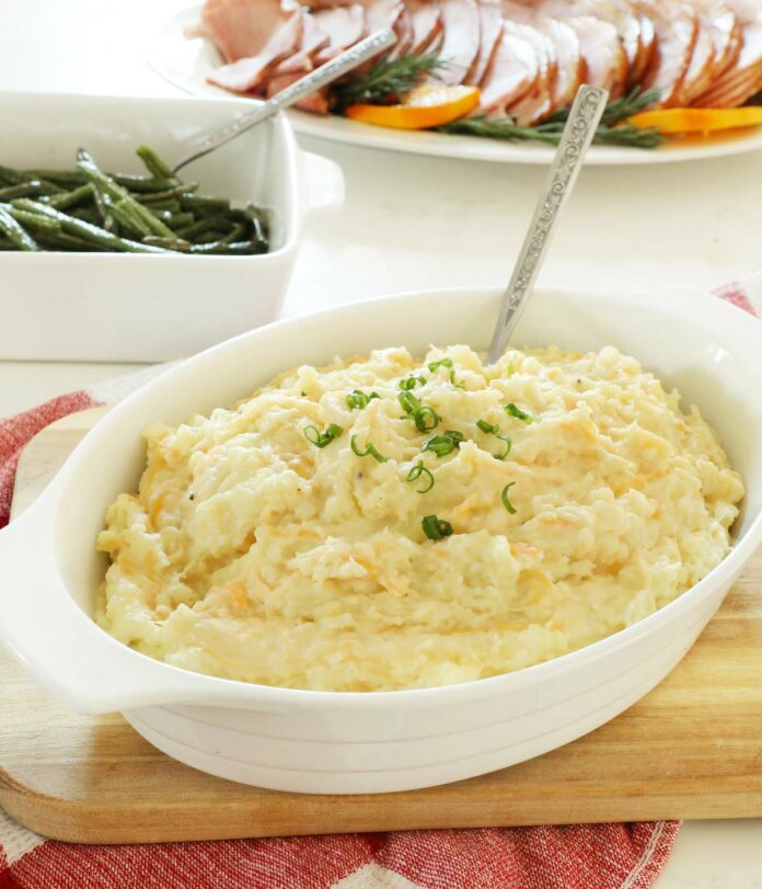 Cheesy mashed potatoes in a white serving dish on a wooden cutting board.